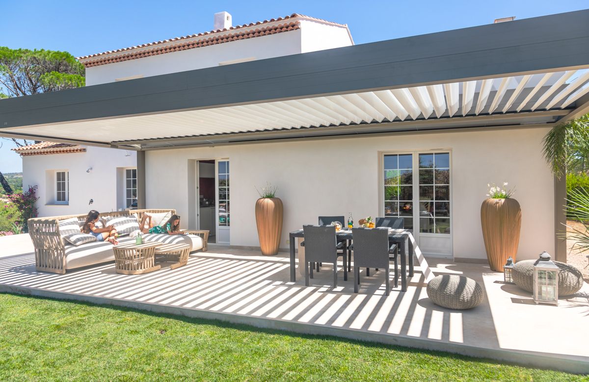 Terrasse moderne avec pergola bioclimatique grise adossée à une maison contemporaine, salon de jardin et espace repas extérieur ensoleillé.